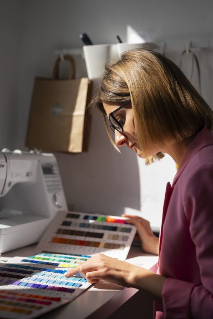 Side view of a woman selecting thread colors from an embroidery color chart beside a sewing machine, in natural daylight